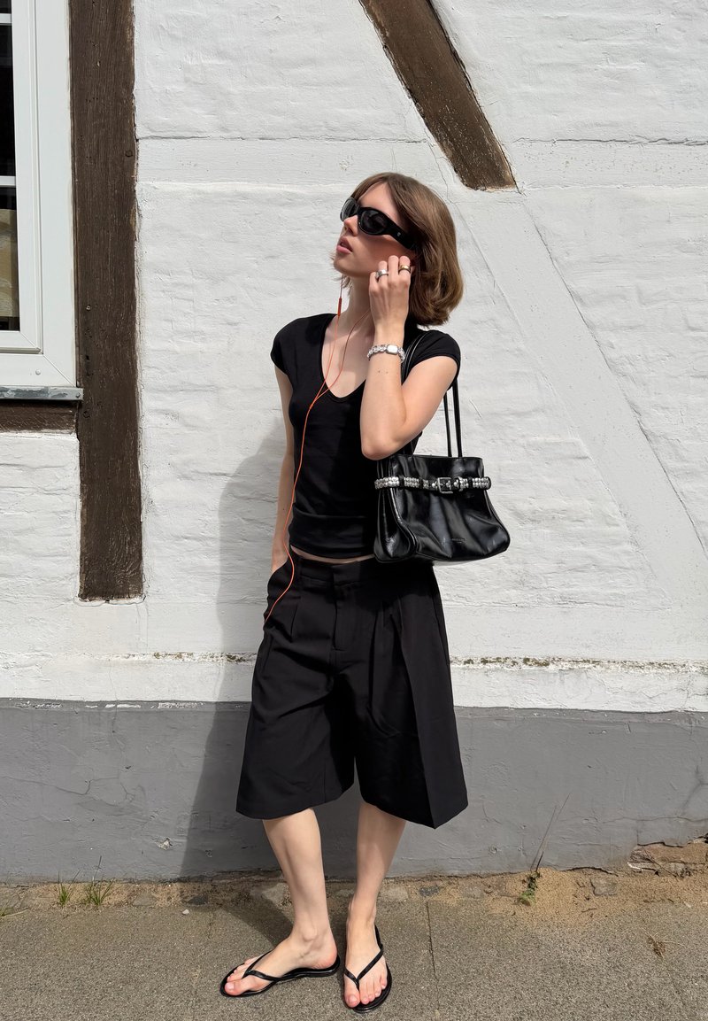 Jeune femme en tenue noire avec lunettes de soleil et sac à main, debout contre un mur blanc, ajustant des écouteurs connectés à un appareil.