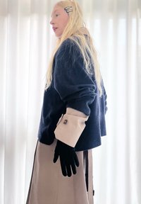 Blonde woman with hair clip wears navy jumper with beige buttoned wide cuffs, beige skirt, and black gloves, standing against white curtains.
