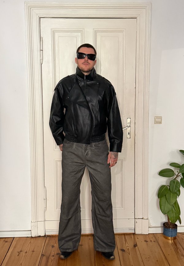 Homme portant une veste en cuir noire, des lunettes de soleil foncées et un pantalon gris, debout à l'intérieur contre une porte blanche fermée avec un sol en bois et une plante à proximité.