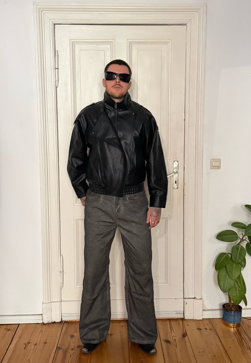 Man wearing black leather jacket, dark sunglasses, and grey pants standing indoors against a closed white door with wooden floor and plant nearby.