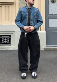 Modrá džínová bunda s límečkem, černá kravata, volné černé kalhoty a sportovní boty s bílými a šedými akcenty. Klíče připevněné na pásku.