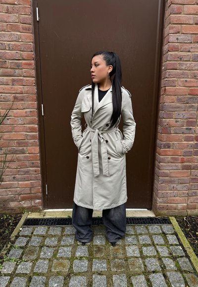 Trench coat gris claro con cintura ajustada, botones de doble botonadura y hombreras. Pantalones de mezclilla oscuros de corte oversized y una camiseta negra debajo.