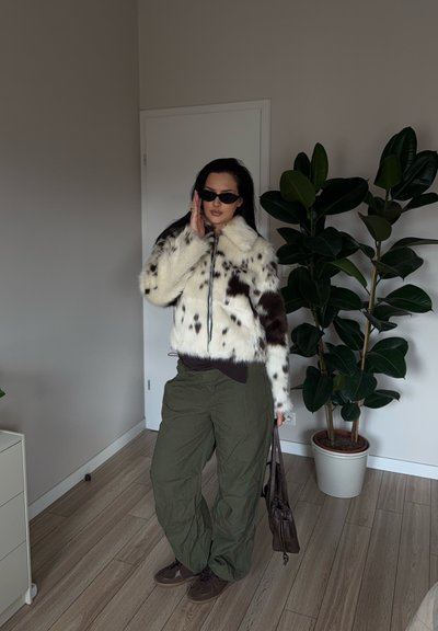 Chaqueta de piel sintética blanca con manchas marrones, de diseño corto, combinada con pantalones cargo verdes oscuros y zapatos marrones, sosteniendo un bolso marrón.