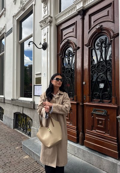 Un abrigo trinchera beige, pantalones negros y gafas de sol oscuras combinados con un bolso texturizado marrón claro. Puertas de madera elegantes con trabajos en metal ornamentales.