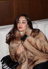 Cappotto in pelliccia sintetica in tonalità di marrone con una texture morbida; il modello indossa guanti neri, seduto su un divano bianco, con una piccola borsa nera.