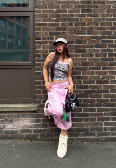 Bolso negro, pantalones de chándal rosa claro, top sin mangas gris con patrones, botas gruesas blancas, cabello largo con reflejos de colores, frente a una pared de ladrillo.