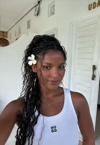 Femme aux longs cheveux tressés avec une fleur blanche, portant un débardeur blanc Hugo et un collier en or, debout à l'intérieur devant des murs et une porte blancs.