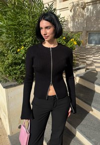 Γυναίκα με κοντό μαύρο καρέ χτένισμα, που φοράει μαύρο ριμπ πλεκτό πουλόβερ με φερμουάρ και παντελόνια, κρατώντας ροζ τσάντα, στέκεται κοντά σε φυτά και σκάλες κάτω από το ηλιακό φως