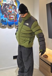 Άτομο που φοράει πράσινο και γκρι μπουφάν North Face, μαύρο σκουφάκι με λευκό λογότυπο και μαύρο παντελόνι, στέκεται μέσα σε εσωτερικό χώρο κοντά σε τέχνη με σκέιτμπορντ.