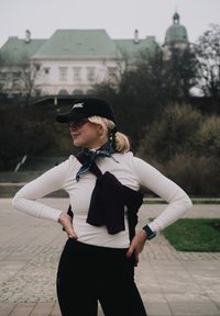 Witte top met lange mouwen, paarse trui om de taille gebonden, zwarte legging en een zwarte pet. Accessoires zijn onder andere een zonnebril en een sjaal met patroon.