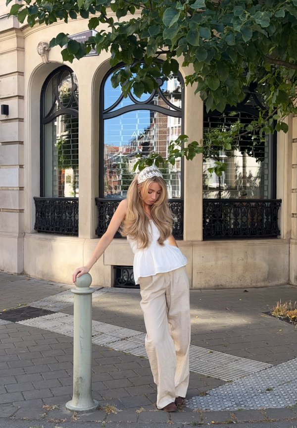 Donna con lunghi capelli biondi e fascia di pizzo si appoggia a un palo basso sul marciapiede in città vicino a un edificio con finestre ad arco e verde.