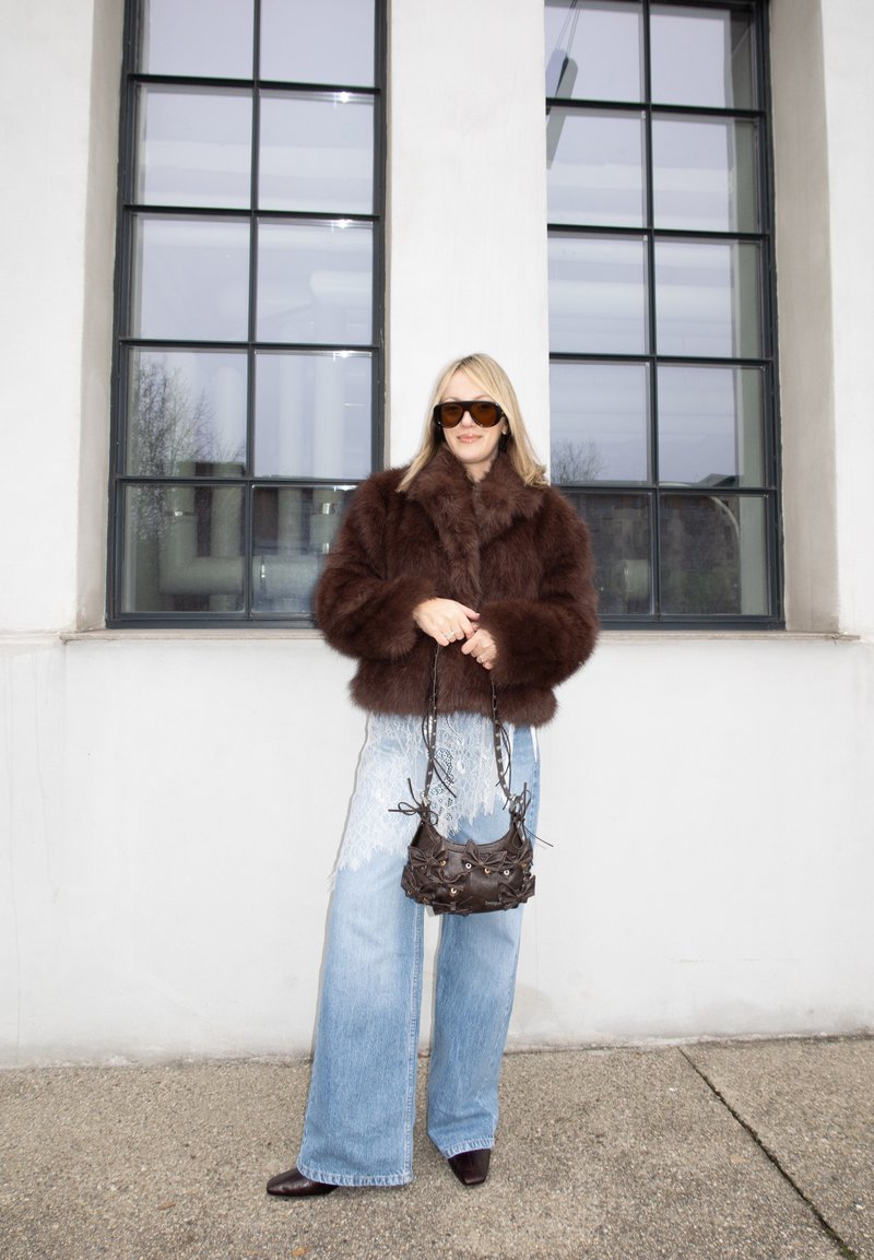 Femme portant une veste en fourrure brune, lunettes de soleil surdimensionnées, jeans larges avec des détails en dentelle, tenant un petit sac à main marron, debout près d'une grande fenêtre.