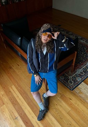 Conjunto informal que incluye un cárdigan a rayas negro y azul sobre una camisa blanca, pantalones cortos de mezclilla y zapatillas negras. Los accesorios incluyen gafas de sol.
