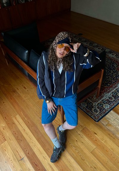 Conjunto informal que incluye un cárdigan a rayas negro y azul sobre una camisa blanca, pantalones cortos de mezclilla y zapatillas negras. Los accesorios incluyen gafas de sol.