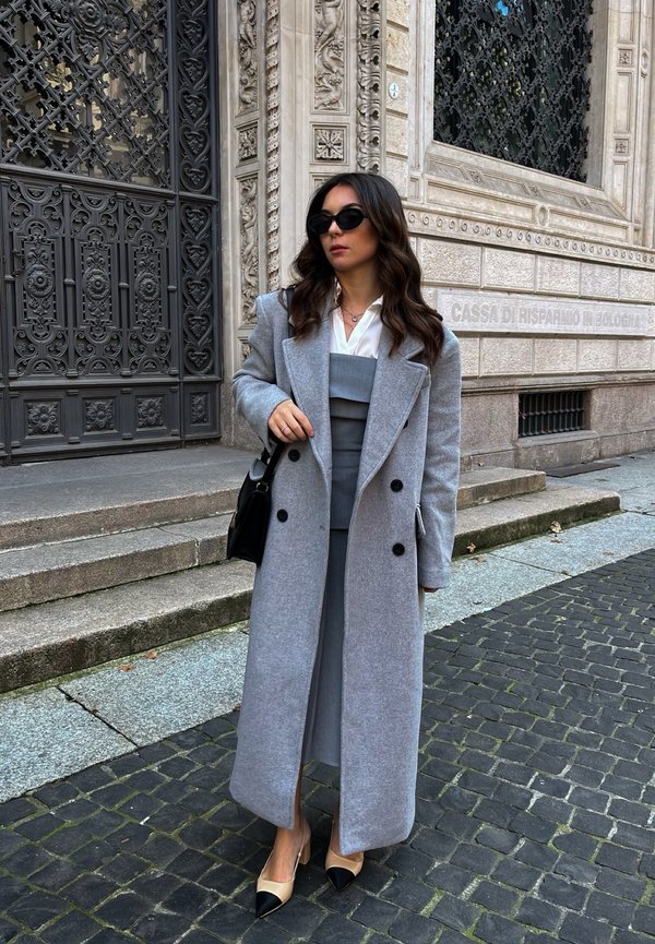 Grijze wollen jas met grote zwarte knopen, gelaagd over een grijze jurk. Accessoires zijn een zwarte handtas en beige hakken met zwarte neus.Â 