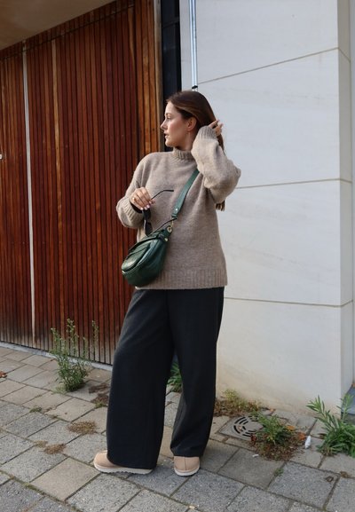 Suéter de punto beige, pantalones anchos negros y zapatillas beige. Bolso de hombro verde con diseño fruncido. Entorno al aire libre con fondo de madera.