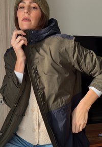 Femme portant un bonnet en tricot, superposée d'un gilet en polaire beige et d'une veste zippée bicolore, ajustant le col de la veste à l'intérieur.