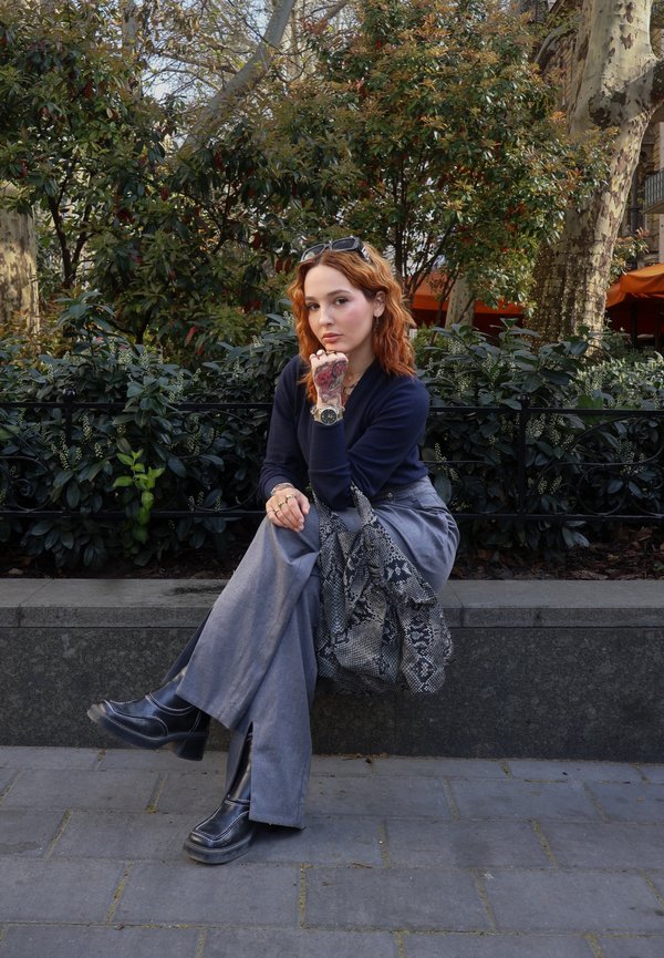 Jeune femme aux cheveux roux assise sur un banc en pierre, portant un haut bleu marine, un pantalon gris, des bottes noires et tenant un sac à motifs à l'extérieur.