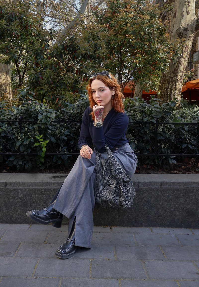 Jeune femme aux cheveux roux assise sur un banc en pierre, portant un haut bleu marine, un pantalon gris, des bottes noires et tenant un sac à motifs à l'extérieur.