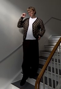 Cardigan a cuadros marrones y beige sobre una camiseta blanca, combinado con pantalones negros holgados. De pie en unas escaleras, sosteniendo gafas, en un ambiente casual.