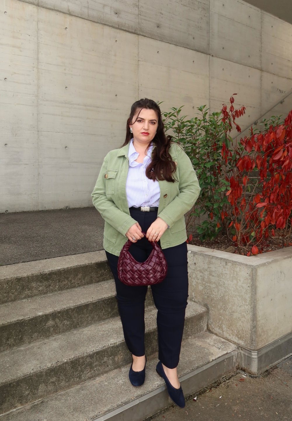 Vrouw die op een buitenbetonnen trap staat, gekleed in een groene jas, gestreept shirt, donkere broek, met een gestructureerde bordeauxrode handtas in de hand, omringd door rode en groene planten.