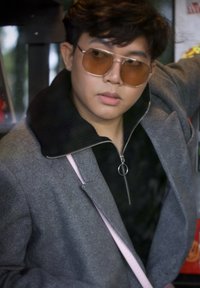 Γκρίζο παλτό από μίγμα μαλλιού με ψηλό γιακά, συνδυασμένο με μαύρο φούτερ με φερμουάρ. Μεγάλες καφέ ηλιόμπαρες και ελαφριά ροζ θήκη ώμου ορατές.