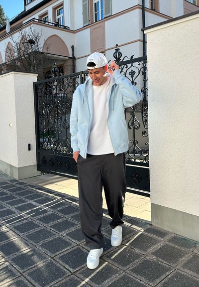 Sudadera de cremallera en azul claro sobre una camiseta blanca, combinada con pantalones negros y zapatillas blancas, de pie frente a una puerta decorativa.