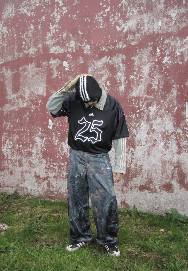 Black sports jersey with number 25, denim shirt underneath, and patterned cargo pants. Gray and purple color scheme; standing on grass.