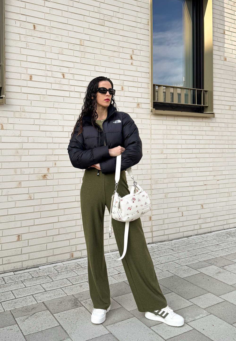 Μαύρο padded μπουφάν, ελαιόχρωμα ριγωτά παντελόνια με φαρδιά μπατζάκια, λευκά αθλητικά παπούτσια με πράσινες λεπτομέρειες και τσάντα με λουλουδάτ�ο σχέδιο.