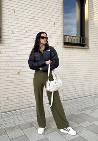 Μαύρο padded μπουφάν, ελαιόχρωμα ριγωτά παντελόνια με φαρδιά μπατζάκια, λευκά αθλητικά παπούτσια με πράσινες λεπτομέρειες και τσάντα με λουλουδάτο σχέδιο.