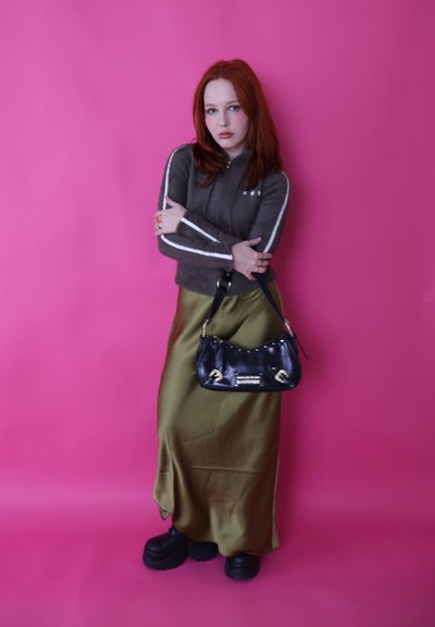 Mujer pelirroja con suéter gris con cremallera, falda de satén verde oliva, zapatos negros de plataforma, sosteniendo un bolso negro contra un fondo rosa brillante.