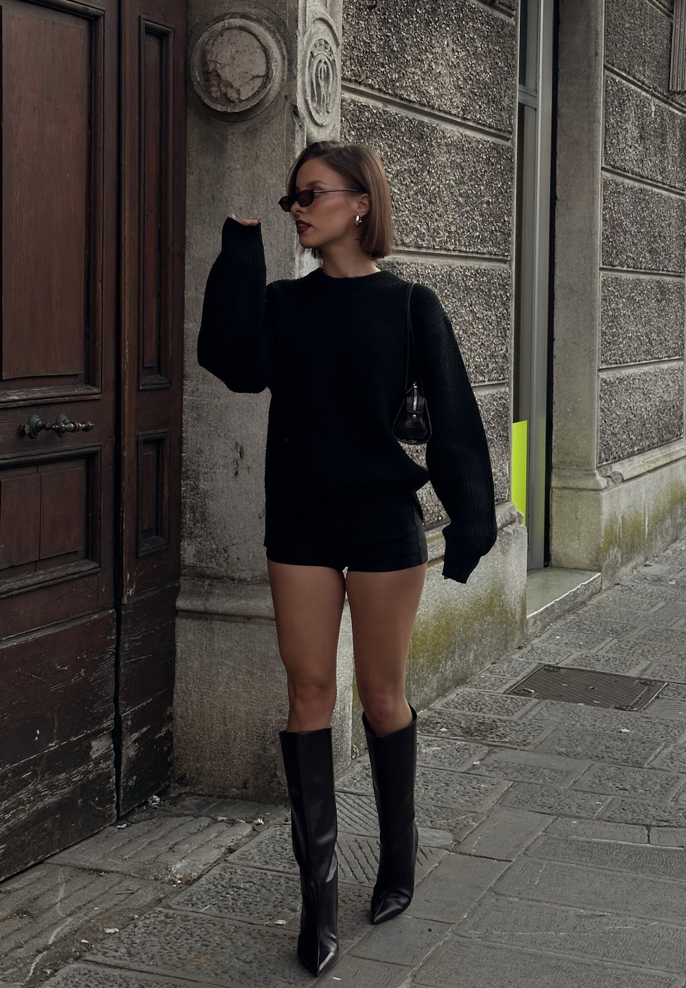 Čierny pletený sveter s nadrozmernými rukávmi, skombinovaný s čiernymi šortkami a čižmami na vysokom opätku po kolená s ostrou špičkou. Ekologická kabelka.