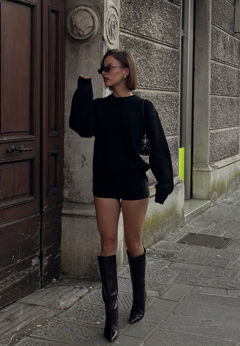 Pulover negru tricotat cu mâneci supradimensionate, asortat cu pantaloni scurți negri și cizme cu toc, vârf ascuțit, până la genunchi. Geantă ecologică.