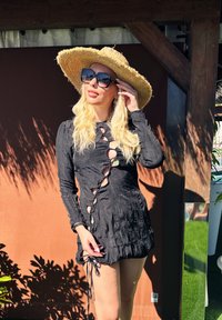 Vestido preto com tecido texturizado, apresentando detalhes recortados, mangas longas e atilhos ajustáveis na bainha. Combinado com um chapéu de palha e óculos de sol.