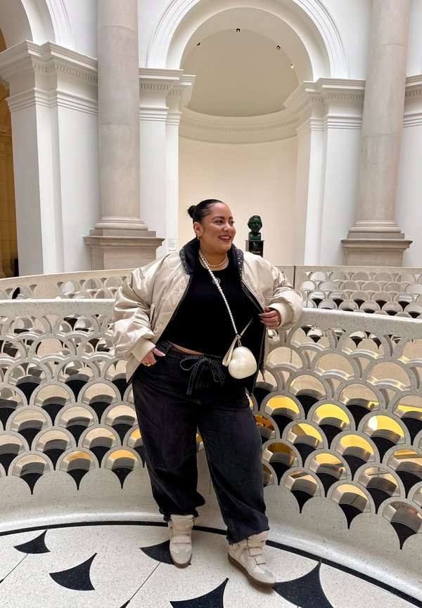 Persoană zâmbitoare, sprijinită de o balustradă ornamentată cu modele arcuite în interiorul unei clădiri cu coloane mari și o statuie bust în fundal.