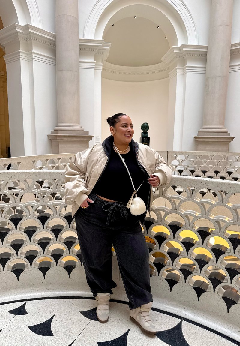 Personne souriante, appuyée sur une rambarde ornée de motifs en arc à l'intérieur d'un bâtiment avec de grandes colonnes et un buste en arrière-plan.
