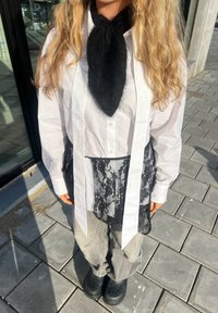 Personne aux longs cheveux blonds portant une chemise blanche, un tablier noir en dentelle, une écharpe noire, un pantalon gris et des chaussures noires sans lacets, debout sur un trottoir carrelé.