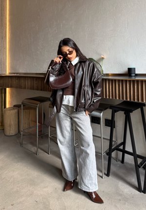 Mujer con chaqueta de cuero marrón oscuro de gran tamaño, camisa blanca, vaqueros anchos gris claro, botas marrones puntiagudas, gafas de sol, sosteniendo un bolso marrón en interiores.