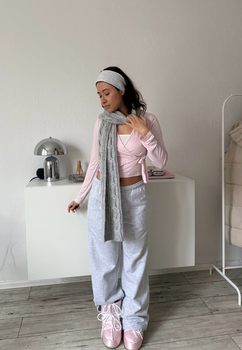 Femme en vêtements décontractés avec une écharpe en maille grise et un bandeau, debout dans une pièce minimaliste avec des meubles blancs et un sol en bois.