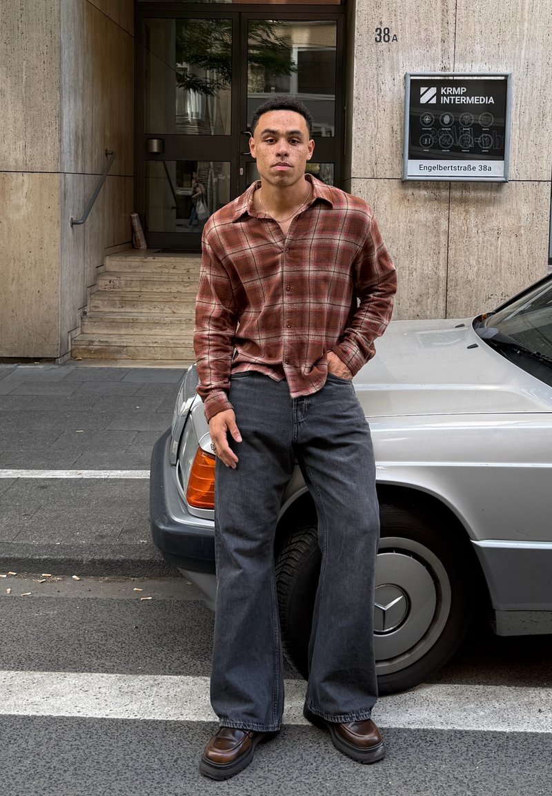 Jeune homme en chemise à carreaux marron et jean noir à jambes larges appuyé contre une voiture argentée garée dans une rue de la ville.