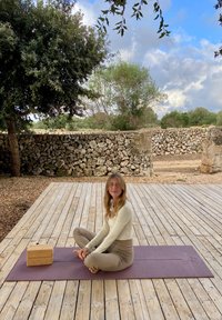 Tapete de yoga roxo, bloco de cortiça leve, superfície de deck de madeira, parede de pedra natural ao fundo, folhagem verde em camadas, céu nublado.