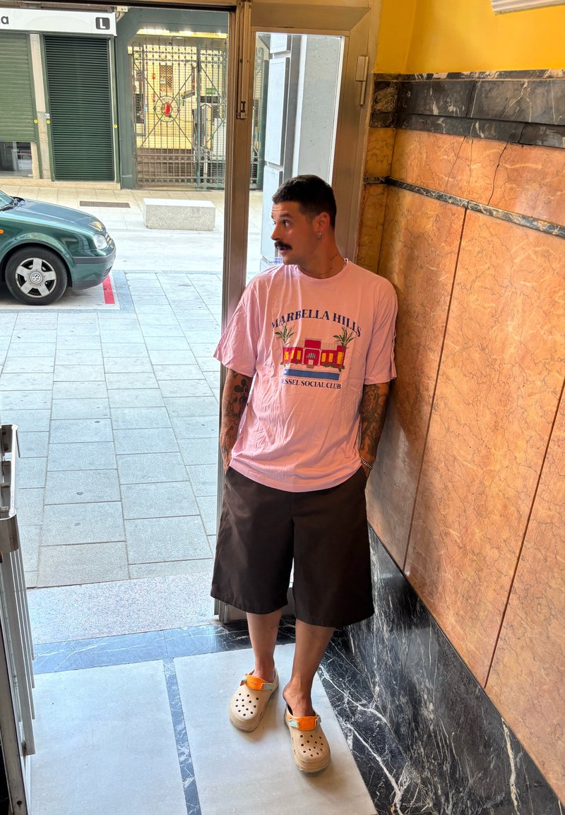 Lichtroze oversized t-shirt met "Marbella Hills" grafiek, bruine shorts en beige klompen met oranje accenten, staand binnen bij een open deur.