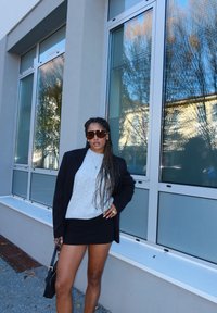 Zwart blazer over een grijze trui, gecombineerd met een zwarte minirok. Bruine zonnebril, gevlochten haar en een gestructureerde zwarte handtas. Stedelijke omgeving.