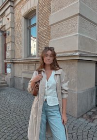 Ανοιχτό μπεζ παλτό πάνω από λευκό πουκάμισο, σε συνδυασμό με ανοιχτό μπλε τζιν. Καφέ τσάντα με λουράκι στον ώμο; στέκεται σε λιθόστρωτο δρόμο.