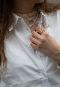 Camisa blanca con cuello y botón, superpuesta sobre una blusa de encaje. Un anillo dorado que asemeja la letra 'M' con gemas incrustadas, llevado en la mano.