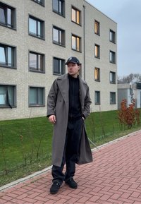 Manteau en laine grise sur un sweat à capuche noir, associé à un pantalon noir et des baskets. En arrière-plan, un chemin en briques et des façades de bâtiments modernes.