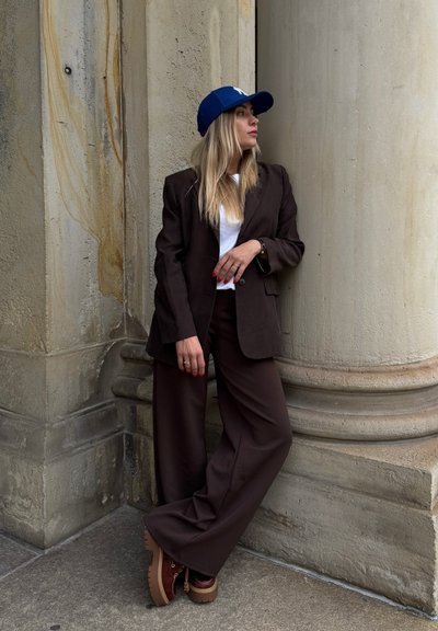 Blazer marrón sobre una camiseta blanca, combinado con pantalones marrones anchos. Una gorra azul y zapatos de plataforma marrones con suelas gruesas completan el look.