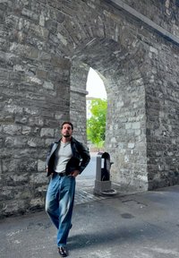 Chaqueta de cuero negra, camiseta henley gris, jeans de pierna ancha azul claro y zapatos oscuros. Fondo de arco de piedra con vegetación visible.