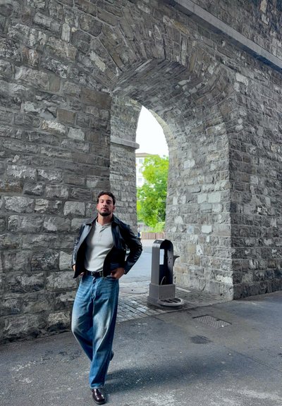 Chaqueta de cuero negra, camiseta henley gris, jeans de pierna ancha azul claro y zapatos oscuros. Fondo de arco de piedra con vegetación visible.