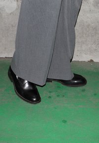 Črni usnjeni loaferji z gladko teksturo, dopolnjeni s sivo črtastimi hlačami, postavljeni na zeleno površino.